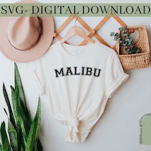 Könnte beinhalten: Ein weißes T-Shirt mit dem Wort "MALIBU" in Schwarz auf der Vorderseite. Das Shirt hängt an einem Holzbügel an einer weißen Wand.