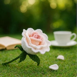Puede incluir: Una rosa de ganchillo hecha a mano con pétalos blancos y centro rosa, tallo y hojas verdes, sobre hierba verde. Una taza y platillo blancos están en el fondo, junto con un libro abierto.