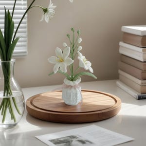 May include: A handmade white crochet vase holds a bouquet of white flowers with yellow centers and green stems. The vase sits on a round wooden tray. A clear glass vase with green stems and white flowers is to the left.