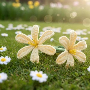 Puede incluir: Dos flores de ganchillo hechas a mano con pétalos de color amarillo pálido y centros amarillos descansan sobre un lecho de hierba verde. Las flores tienen un aspecto delicado y texturizado, y están rodeadas de pequeñas margaritas blancas.
