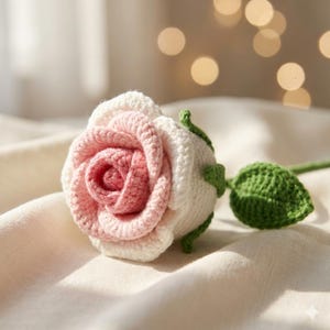 Puede incluir: Una rosa de ganchillo hecha a mano en tonos rosa y blanco, con un tallo y una hoja verdes. Los pétalos de la flor están meticulosamente tejidos a ganchillo, mostrando una textura detallada. La rosa es un encantador artículo decorativo.
