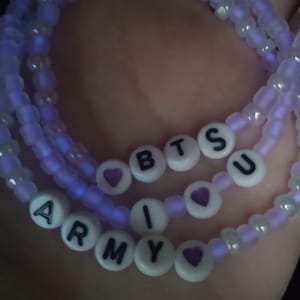 May include: Three beaded bracelets in shades of purple and white. The bracelets have white letter beads spelling out "BTS" and "ARMY" with purple heart accents. The beads glow with a soft, ethereal light.