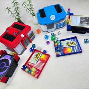 Può includere: Un set di quattro scatole di contenimento per giochi di carte dai colori vivaci con uno schema di colori rosso, blu e bianco. Le scatole hanno una varietà di disegni, tra cui un edificio, una stella e un cerchio. Ci sono anche diversi dadi sparsi intorno alle scatole.