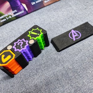 Puede incluir: Un conjunto de piezas de juego coloridas, que incluyen una pieza rectangular negra con un logotipo de los Vengadores morado y un soporte negro con varias fichas de colores. Las fichas presentan signos de interrogación, exclamaciones y otros símbolos.