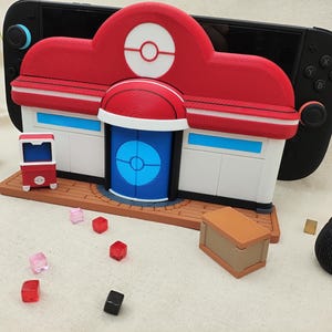 Op de afbeelding: Een rood, wit en blauw 3D-geprint model van een Pokémon Center, met een blauwe deur en een bruine basis. Een kleine rode automaat en een bruine doos zijn ook aanwezig. Een zwarte spelconsole staat op de achtergrond.