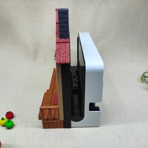 May include: A white and black video game console with a red, white, and brown miniature building attached to the side.