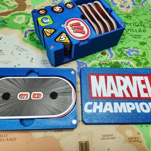 以下が含まれることがあります： 前面に「MARVEL CHAMPIONS」と書かれた青と赤の箱。箱が開いており、青と白のサイコロタワーとサイコロが入っている。サイコロタワーには、異なる数字が書かれたサイコロがいくつかある。サイコロタワーは、青と緑の背景の地図の上に置かれている。