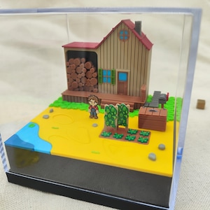 Könnte beinhalten: Miniatur-Diorama-Szene in einer transparenten Acrylbox. Es zeigt ein kleines Holzhaus mit rotem Dach, eine Figur, einen Garten und einen Brunnen. Die Basis ist gelb mit einem blauen Wasserabschnitt. Dekorative Würfel befinden sich im Hintergrund.