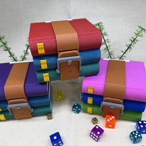 Op de afbeelding: Drie kleurrijke, gestapelde dozen die op boeken lijken, elk vastgezet met een bruine riem en gesp. De dozen zijn rood, paars en roze, met blauwe en groene lagen. Dobbelstenen in verschillende kleuren liggen verspreid.