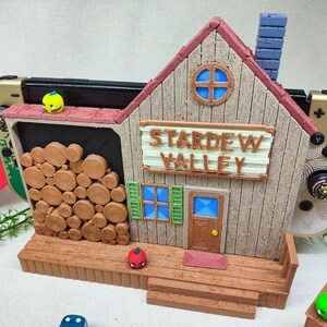 May include: A 3D printed model of the Stardew Valley farmhouse, painted in brown and beige with a red roof. The model is designed to hold a Nintendo Switch console and has a wood-like texture. The sign above the door reads "Stardew Valley".