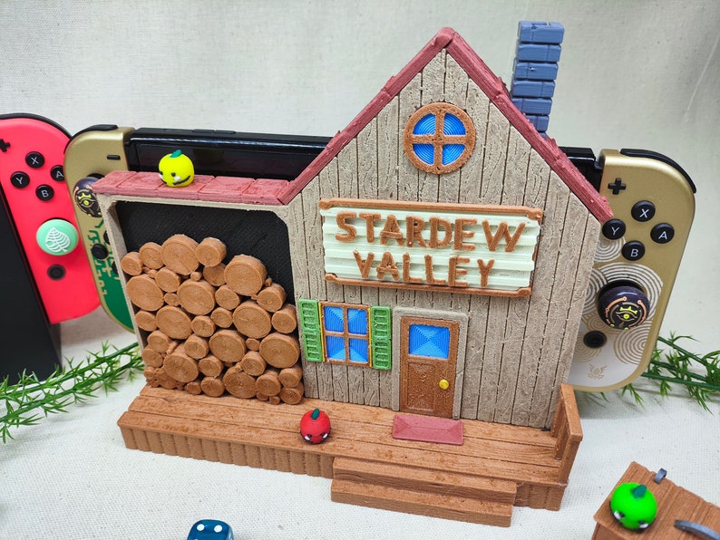May include: A 3D printed model of the Stardew Valley farmhouse, painted in brown and beige with a red roof. The model is designed to hold a Nintendo Switch console and has a wood-like texture. The sign above the door reads "Stardew Valley".