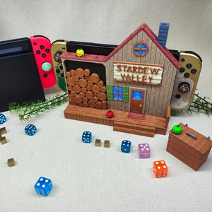 May include: A wooden Stardew Valley themed video game console stand with two Nintendo Switch controllers, a stack of logs, and a small green box with a green apple on top. There are also several blue and orange dice scattered around the stand.