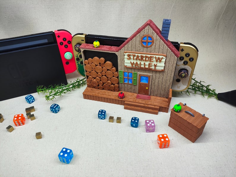 May include: A wooden Stardew Valley themed video game console stand with two Nintendo Switch controllers, a stack of logs, and a small green box with a green apple on top. There are also several blue and orange dice scattered around the stand.