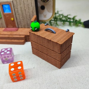 May include: A wooden box with a hinged lid and a metal handle. The box is painted brown and has a textured surface. There are three dice on the table next to the box, one blue, one purple, and one orange.