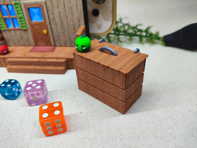 May include: A wooden box with a hinged lid and a metal handle. The box is painted brown and has a textured surface. There are three dice on the table next to the box, one blue, one purple, and one orange.