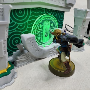 Peut inclure: Figurine détaillée d'un personnage avec un arc et des flèches, sur fond d'emblème circulaire vert brillant et de structures architecturales. La scène est éclairée d'une lumière verte, suggérant un thème fantastique ou de jeu vidéo.