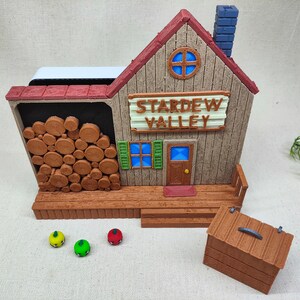 May include: A brown wooden model of the Stardew Valley shop with a red roof, a blue chimney, and a sign that reads "Stardew Valley". The model has a wooden porch with a small wooden chest in front of it.