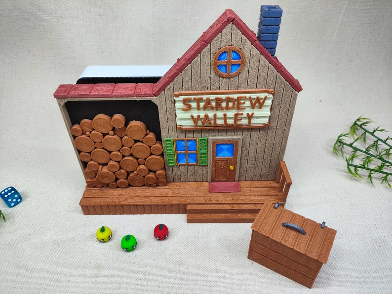 May include: A brown wooden model of the Stardew Valley shop with a red roof, a blue chimney, and a sign that reads "Stardew Valley". The model has a wooden porch with a small wooden chest in front of it.