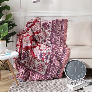 May include: A red, white and blue patterned throw blanket with fringe on the edges. The blanket is draped over a sofa with a white cushion.