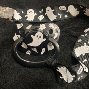 Puede incluir: Un chupete negro y blanco con un dise&ntilde;o de fantasma y una cinta negra y blanca con dise&ntilde;os de fantasmas y "boo". La cinta tiene un clip de pl&aacute;stico transparente.