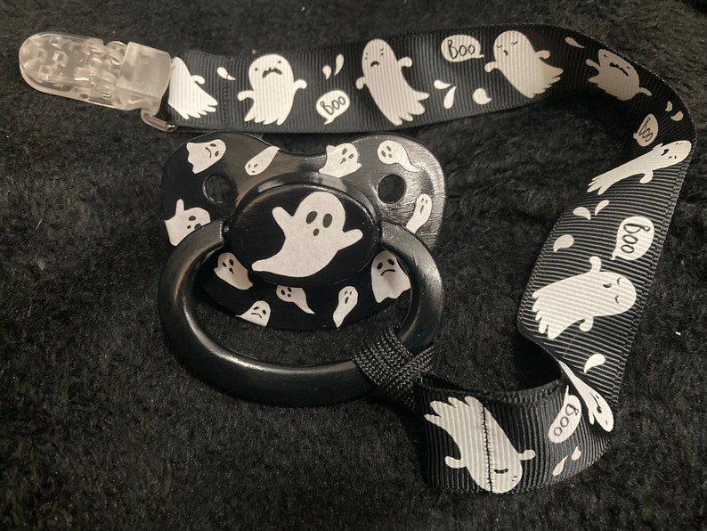 Puede incluir: Un chupete negro y blanco con un dise&ntilde;o de fantasma y una cinta negra y blanca con dise&ntilde;os de fantasmas y "boo". La cinta tiene un clip de pl&aacute;stico transparente.
