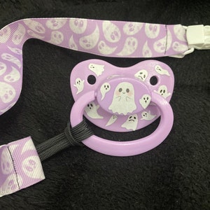 Puede incluir: Un chupete morado con un dise&ntilde;o de fantasma blanco. El chupete tiene un clip unido a una cinta morada y blanca con un patr&oacute;n de fantasma.