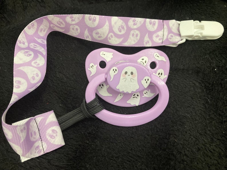 Puede incluir: Un chupete morado con un dise&ntilde;o de fantasma blanco. El chupete tiene un clip unido a una cinta morada y blanca con un patr&oacute;n de fantasma.