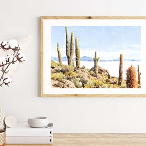 May include: Watercolor painting of a desert landscape with tall cacti and a body of water in the background. The painting is framed in a light wood frame.