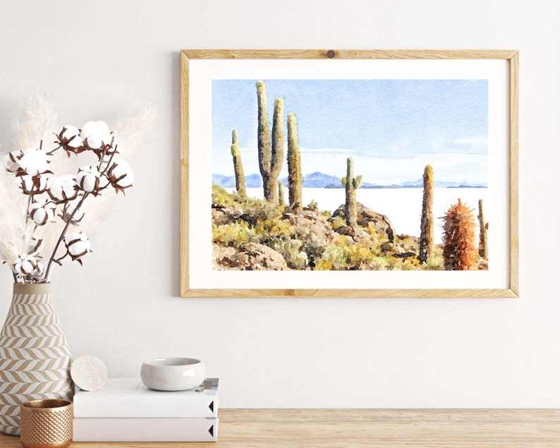 May include: Watercolor painting of a desert landscape with tall cacti and a body of water in the background. The painting is framed in a light wood frame.