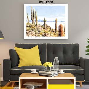 May include: A watercolor painting of a desert landscape with tall cacti and a body of water in the background. The painting is framed and hanging on a wall in a living room setting.