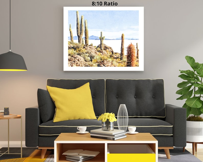 May include: A watercolor painting of a desert landscape with tall cacti and a body of water in the background. The painting is framed and hanging on a wall in a living room setting.