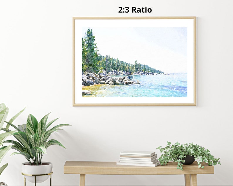 May include: A watercolor painting of a lake scene with a shoreline of trees and rocks. The water is a light blue color and the sky is a light blue color. The painting is framed in a light wood frame.