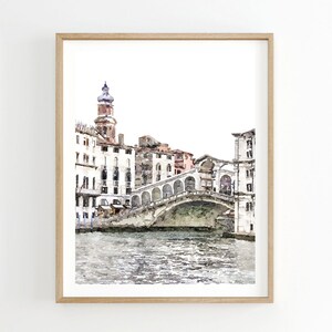 Puede incluir: Pintura a la acuarela del Puente de Rialto en Venecia, enmarcada en madera clara. La obra representa el puente sobre un canal, con edificios bordeando la orilla del agua. La paleta de colores es suave, con tonos apagados de gris, blanco y marr&oacute;n.