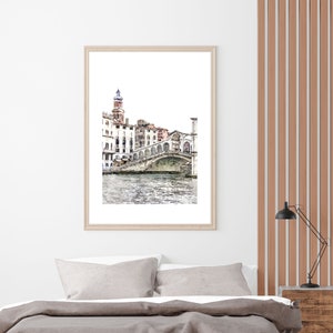 Puede incluir: Una impresi&oacute;n en acuarela enmarcada de un puente veneciano sobre un canal. La obra de arte presenta un marco de madera de color claro y representa edificios y el puente en tonos suaves y apagados. La impresi&oacute;n se muestra encima de una cama con ropa de cama gris.