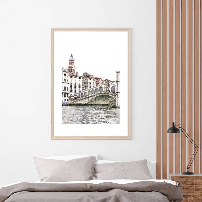 Puede incluir: Una impresi&oacute;n en acuarela enmarcada de un puente veneciano sobre un canal. La obra de arte presenta un marco de madera de color claro y representa edificios y el puente en tonos suaves y apagados. La impresi&oacute;n se muestra encima de una cama con ropa de cama gris.