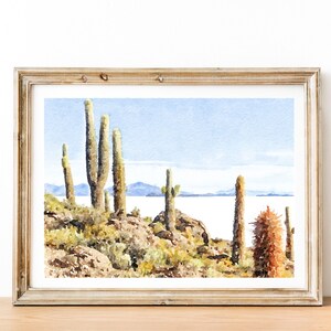 May include: A watercolor painting of a desert landscape with tall cacti and a body of water in the background. The painting is framed in a light wood frame.