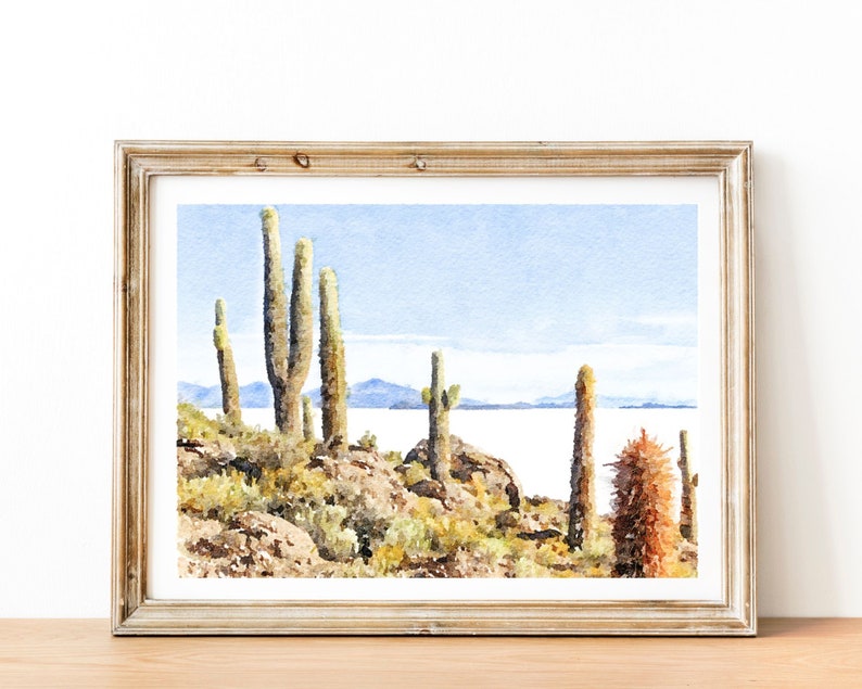 May include: A watercolor painting of a desert landscape with tall cacti and a body of water in the background. The painting is framed in a light wood frame.