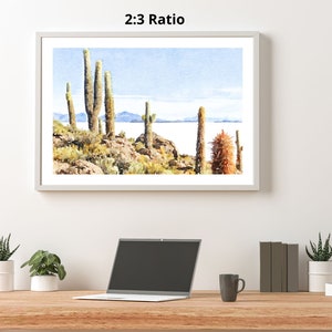May include: A watercolor painting of a desert landscape with tall cacti and a body of water in the background. The sky is a light blue with white clouds. The painting is framed in a white frame.