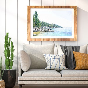 May include: A framed watercolor painting of a lake scene with trees and rocks along the shoreline. The painting is in a light wood frame and is hanging on a white wall above a gray couch with pillows.