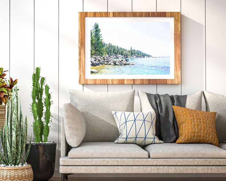 May include: A framed watercolor painting of a lake scene with trees and rocks along the shoreline. The painting is in a light wood frame and is hanging on a white wall above a gray couch with pillows.