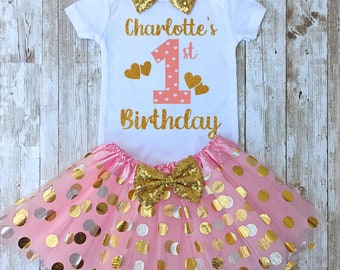 May include: A white short-sleeved bodysuit with the text Charlotte's 1st Birthday in gold glitter, a pink number one, and gold hearts. It includes a pink tulle tutu skirt with gold and silver polka dots and a gold sequin bow headband.