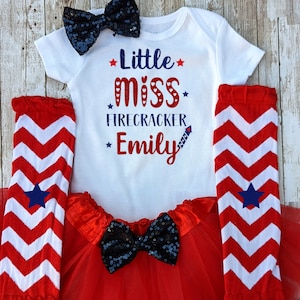 May include: A white onesie with red, white, and blue stars and text that reads "Little miss firecracker Emily". The onesie is paired with a red tulle skirt, a black sequined bow, and red and white chevron leg warmers with blue stars.
