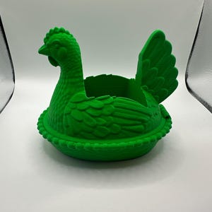 May include: A vivid green hen-shaped container. The container is designed to look like a hen in a nest. The hen has a detailed comb, beak, and feather texture. The nest has a beaded edge. The container is made of a solid, opaque material.