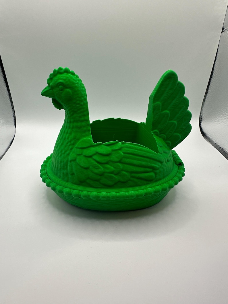 May include: A vivid green hen-shaped container. The container is designed to look like a hen in a nest. The hen has a detailed comb, beak, and feather texture. The nest has a beaded edge. The container is made of a solid, opaque material.