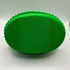 May include: A vibrant green, oval-shaped 3D-printed item. It features a textured surface with horizontal lines and a beaded border. The design suggests a decorative container or tray, with a focus on the detailed edge.