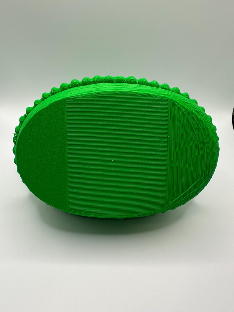 May include: A vibrant green, oval-shaped 3D-printed item. It features a textured surface with horizontal lines and a beaded border. The design suggests a decorative container or tray, with a focus on the detailed edge.