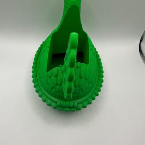May include: A vibrant green 3D-printed dinosaur-shaped container. The container features a textured surface, a long neck, and a handle. The base is round with a decorative edge. It is designed to hold small items.