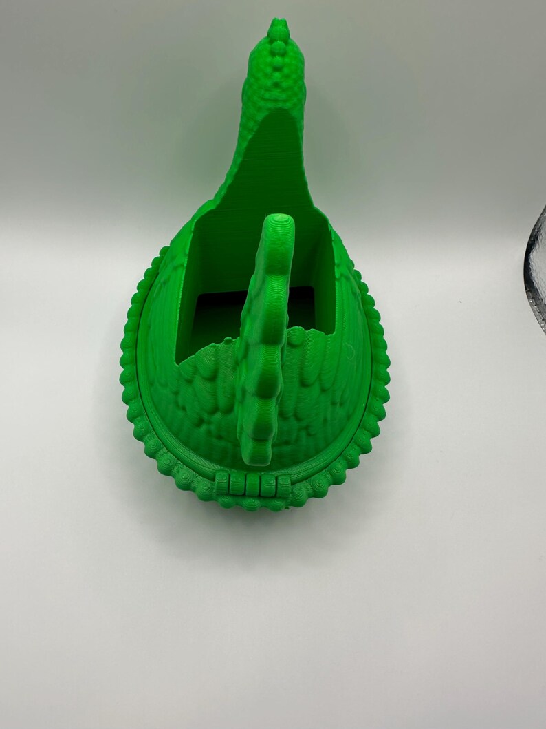 May include: A vibrant green 3D-printed dinosaur-shaped container. The container features a textured surface, a long neck, and a handle. The base is round with a decorative edge. It is designed to hold small items.