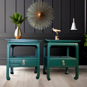 May include: Two green end tables with a single drawer and gold hardware. The tables have a traditional Asian design with curved legs. The tables are set against a black wall with a gold bird figurine and a gold vase with green plants.