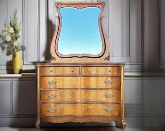 May include: A vintage wooden dresser with a beveled mirror and ornate carvings. The dresser has five drawers with brass hardware and a curved front.